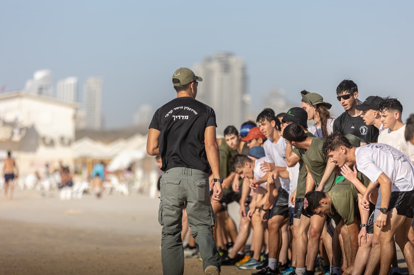 מדריך של אדרנלין כושר קרבי, עם הגב למצלמה, עומד מול שורה ארוכה של חניכים רכונים קדימה על חול החוף