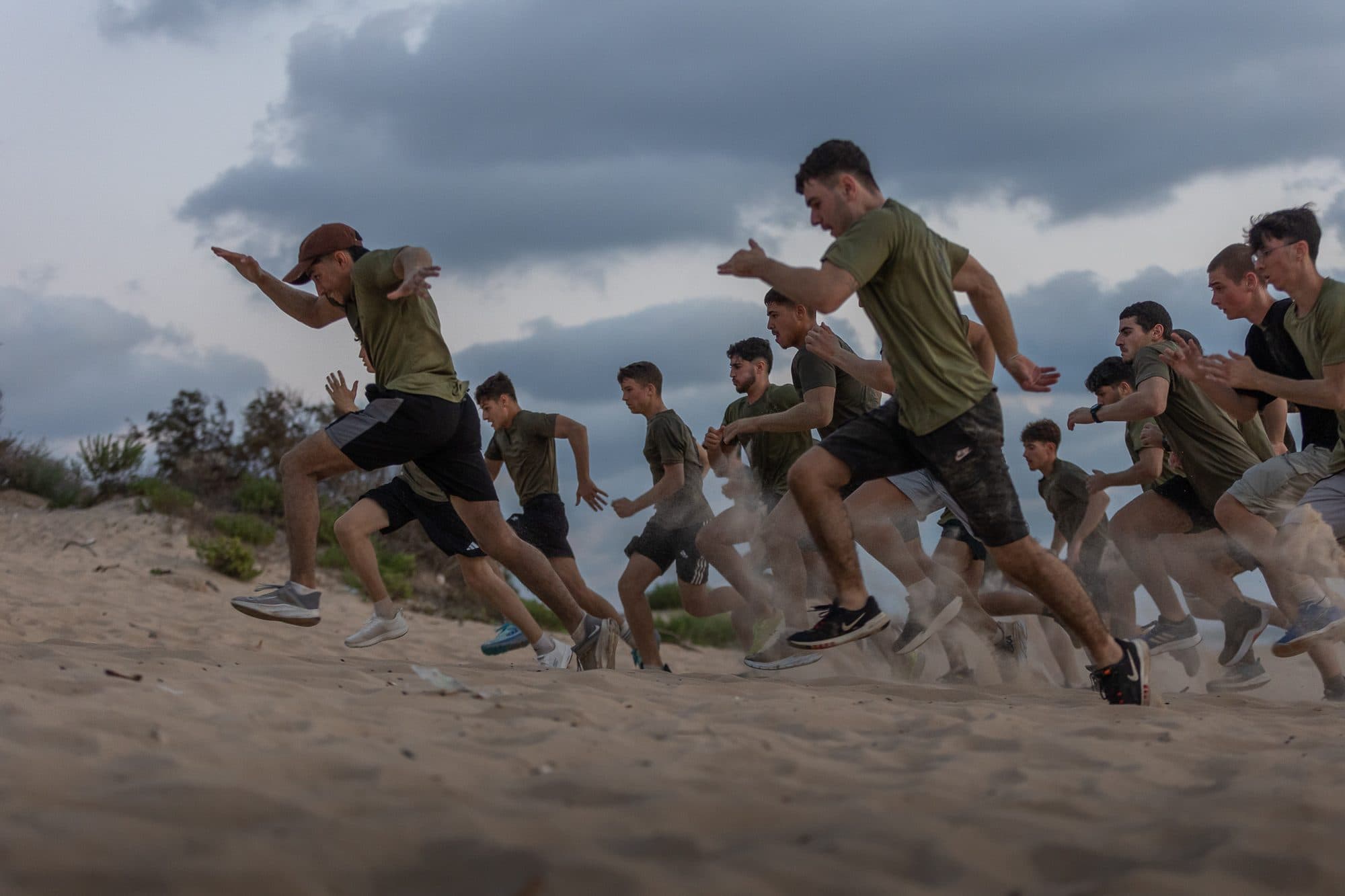 שורה של חניכי אדרנלין כושר קרבי מזנקת בריצה על החול, מתחת לשמיים מעוננים