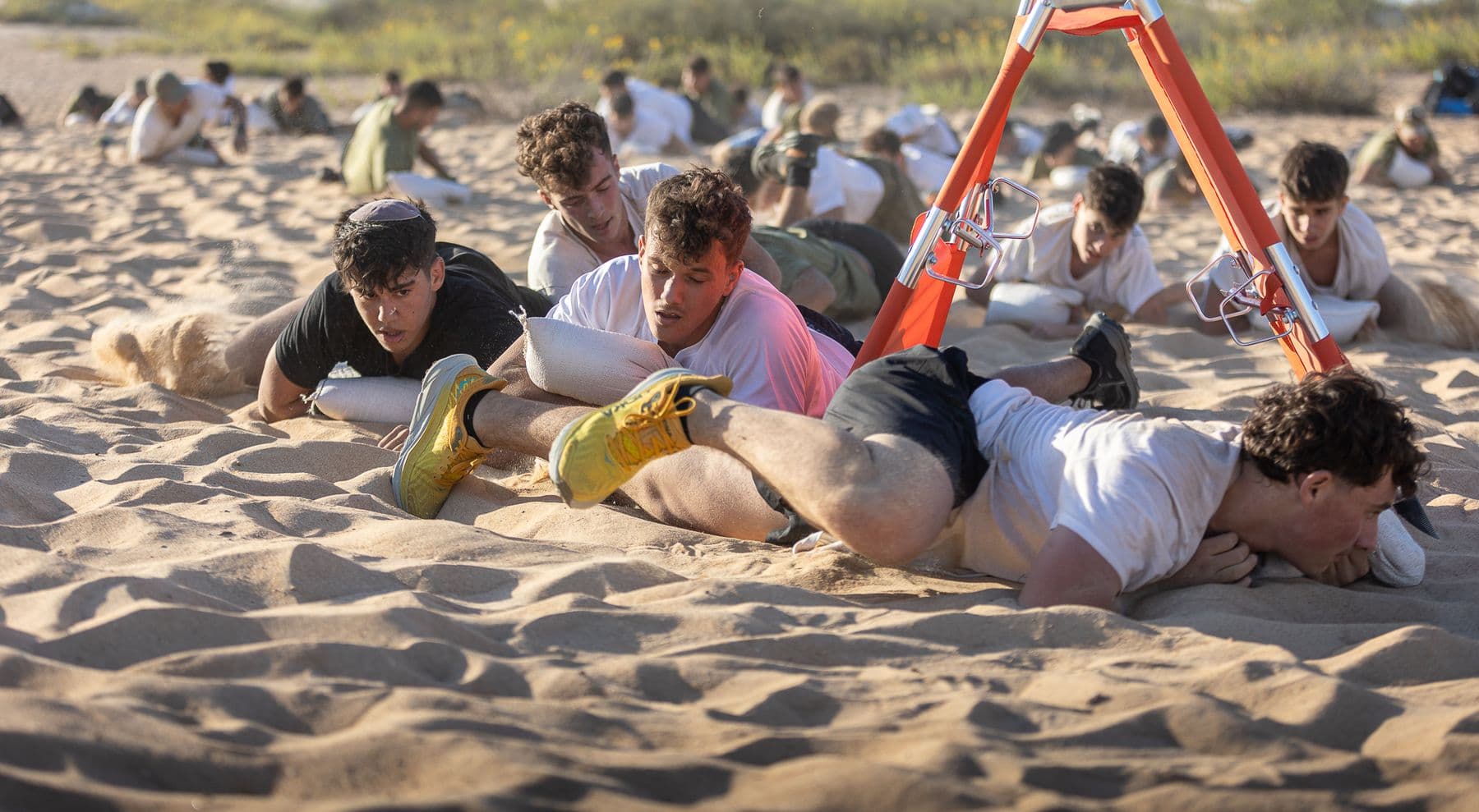חניכי אדרנלין כושר קרבי זוחלים בחול כשכל אחד מהם דוחף לפניו שק, לצד אלונקה כתומה